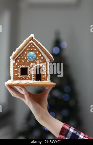 Vertikales Foto. Lebkuchenhaus zu Weihnachten auf Frauenhand auf Weihnachtsbaum hellen Hintergrund. Neujahrsstimmung zu Hause. Geschenke vorbereiten für Stockfoto