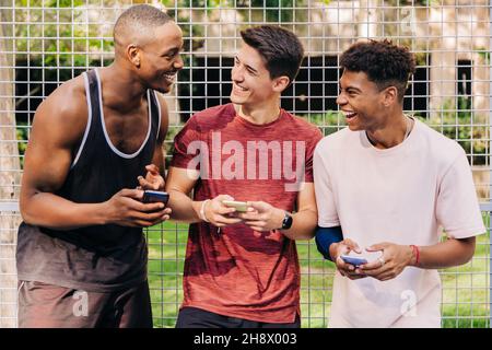 Fröhliche junge, multirassische männliche Athleten in Sportkleidung, die Smartphones surfen und lachen, während sie in der Nähe des Netztors im Stadion stehen Stockfoto