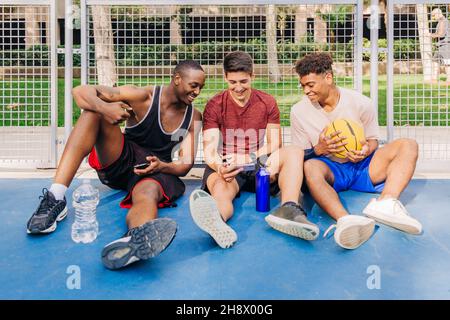 Fröhliche junge, multirassische männliche Athleten in Sportkleidung, die Smartphones durchsuchen und lachen, während sie in der Nähe des Netztors auf dem Sportplatz sitzen Stockfoto
