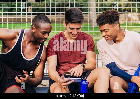 Fröhliche junge, multirassische männliche Athleten in Sportkleidung, die Smartphones durchsuchen und lachen, während sie in der Nähe des Netztors auf dem Sportplatz sitzen Stockfoto