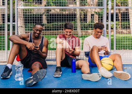 Fröhliche junge, multirassische männliche Athleten in Sportkleidung, die Smartphones durchsuchen und lachen, während sie in der Nähe des Netztors auf dem Sportplatz sitzen Stockfoto