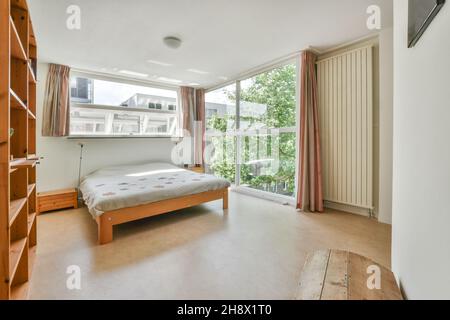 Komfortables Bett mit grauer Decke in der Nähe von Fenstern mit Vorhängen im hellen geräumigen Schlafzimmer mit leeren Holzregalen in der Wohnung platziert Stockfoto