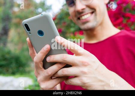 Crop lächelnder junger Mann in stilvollen Brillen lesen gute Nachrichten auf Handy, während im Park mit blühenden Büschen stehen Stockfoto
