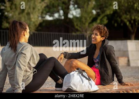 Seitenansicht einer fröhlichen hispanischen Sportlerin im aktiven Ohr, die tagsüber auf dem Boden sitzt und mit einem Freund im Park spricht Stockfoto