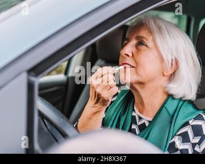 Ältere Frau in eleganter Kleidung, die Lippenstift in den Rückspiegel trägt, während sie auf dem Fahrersitz eines modernen Autos sitzt Stockfoto