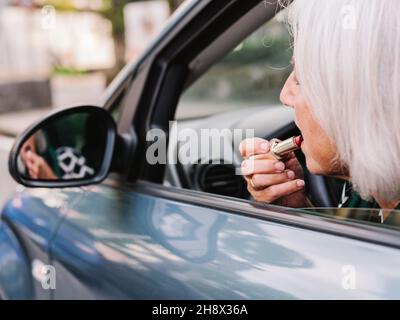 Seitenansicht Ältere Frau in elegantem Stil, die Lippenstift in den Rückspiegel trägt, während sie auf dem Fahrersitz eines modernen Autos sitzt Stockfoto