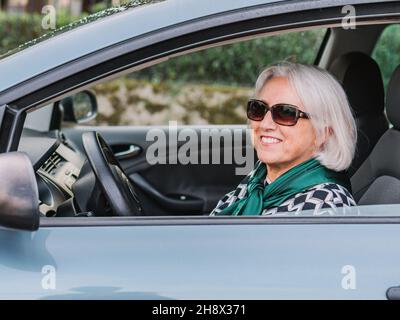 Fröhliche ältere Frau in elegantem Outfit sitzt in einem modernen glänzenden Auto an sonnigen Tagen und schaut weg Stockfoto