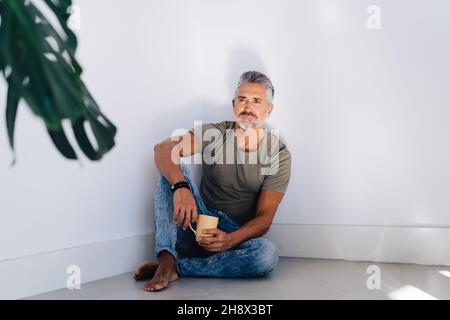 Nachdenklicher barfuß alter Mann in legerer Kleidung, der auf dem Boden in der Ecke sitzt und wegschaut, während er ein heißes Getränk trinkt Stockfoto