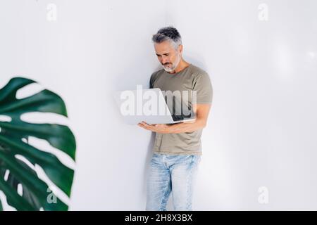 Älterer bärtiger Mann, der den Bildschirm aufmerksam ansieht, während er gegen eine weiße Wand steht und in einem hellen Raum in der Hand einen Laptop durchstöbert Stockfoto