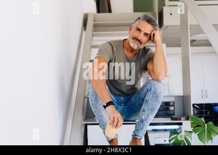 Älterer, fröhlicher Barfußmann, der auf einer Leiter sitzt und die Kamera anschaut, während er sich in einer hellen Wohnung an der Hand lehnt Stockfoto