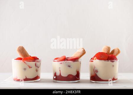 Drei Portionen geschichtetes Dessert in Gläsern mit Cookie Savoyardi, Mascarpone-Käse und Schlagsahne verziert Erdbeeren auf weißem Hintergrund. Stockfoto