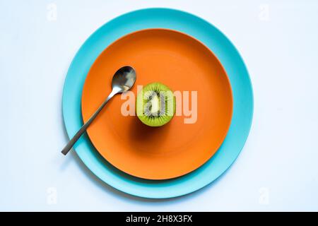 Von oben die Hälfte der frischen reifen saftigen Kiwi und Löffel auf orange und blaue Teller auf weiße Oberfläche gelegt Stockfoto