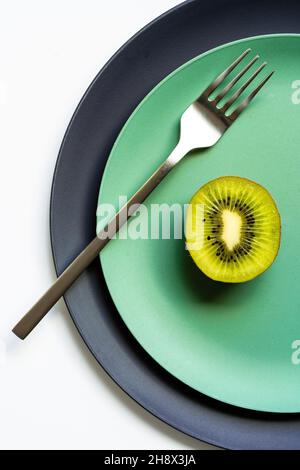 Von oben die Hälfte der frischen reifen saftigen Kiwi und Gabel auf grauen und grünen Tellern auf weißer Oberfläche gelegt Stockfoto