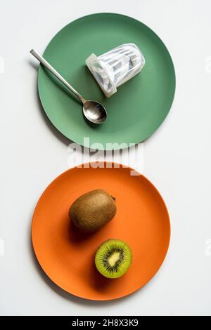 Draufsicht auf frische reife ganze und halbierte Kiwis und Joghurt mit Löffel auf bunte Keramikplatten auf weißem Tisch gelegt Stockfoto