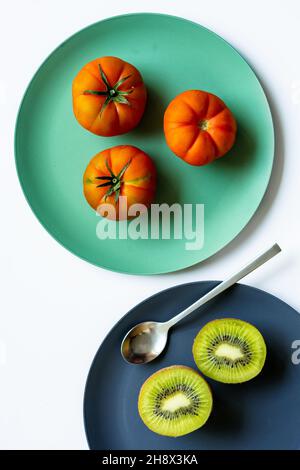 Draufsicht auf frische, gesunde halbierte Kiwi und ganze Tomaten, die auf blauen und türkisfarbenen runden Tellern mit Löffel auf weißem Hintergrund platziert sind Stockfoto