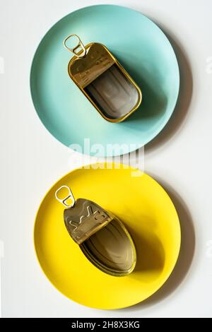 Draufsicht auf rechteckige und ovale geöffnete leere Metalldosen, die auf gelben und blauen Platten auf einem weißen Tisch platziert sind Stockfoto