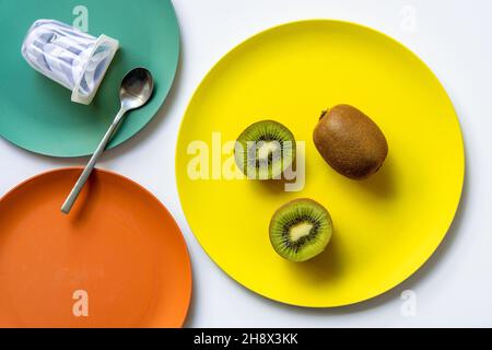 Draufsicht auf frische reife ganze und halbierte Kiwis und Joghurt mit Löffel auf bunte Keramikplatten auf weißem Tisch gelegt Stockfoto