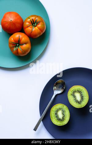 Draufsicht auf frische, gesunde halbierte Kiwi und ganze Tomaten, die auf blauen und türkisfarbenen runden Tellern mit Löffel auf weißem Hintergrund platziert sind Stockfoto