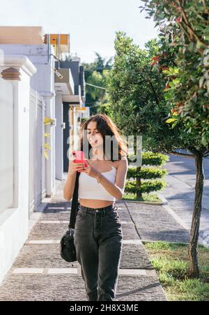 Hispanisch lächelnde Dame, die an sonnigen Sommertagen auf dem Straßenbelag gegen Gebäude und grüne Bäume herumläuft und auf dem Smartphone surft Stockfoto
