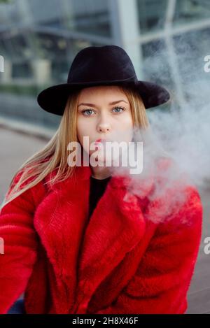 Nette blonde junge Frau mit Hut und roter Jacke, die mit Dampfmaschine auf der Straße raucht Stockfoto