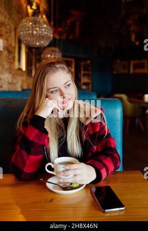 Selbstbewusste, hübsche blonde junge Frau, die eine Tasse Tee im Café hält und wegschaut Stockfoto