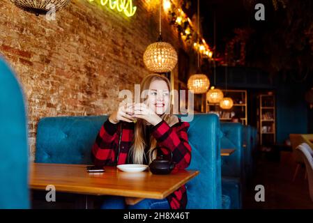 Nette blonde junge Frau, die im Café sitzt und eine Tasse Tee trinkt und wegschaut Stockfoto