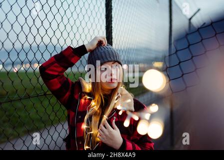Attraktive blonde junge Frau, die sich am Zaun lehnt und eine leichte Girlande aus Glühbirnen hält, die wegschaut Stockfoto