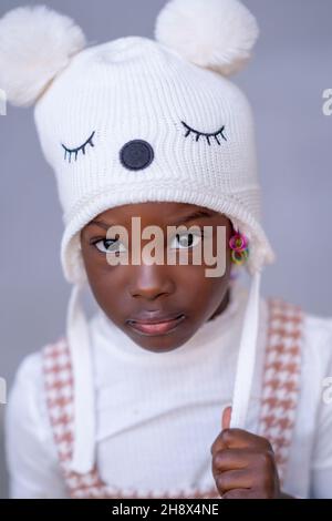 Entzückende afroamerikanische Mädchen Modell in stilvollen warmen Hut mit Pompoms Blick auf die Kamera vor grauem Hintergrund Stockfoto
