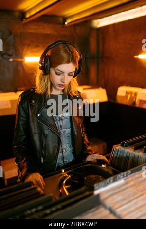 Frau in Lederjacke hört Lied über Vintage-Plattenspieler mit Vinyl-Disc, während sie im Laden steht Stockfoto