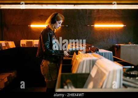 Seitenansicht einer jungen Frau mit Kopfhörern, die Vinyl-Schallplatte auswählt, während sie in der Nähe der Theke mit einer Sammlung von Musikalben steht, die mit glühendem la auf Lager sind Stockfoto