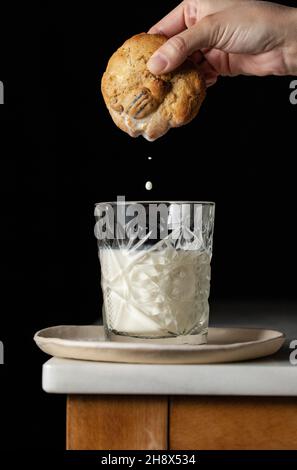 Crop anonyme Person taucht frischen süßen Cookie in ein Glas Milch auf der Ecke des Tisches vor schwarzem Hintergrund in der Küche platziert Stockfoto