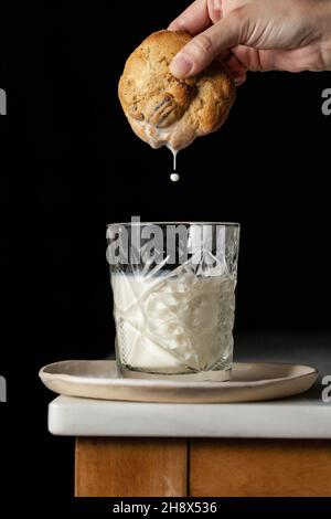 Crop anonyme Person taucht frischen süßen Cookie in ein Glas Milch auf der Ecke des Tisches vor schwarzem Hintergrund in der Küche platziert Stockfoto