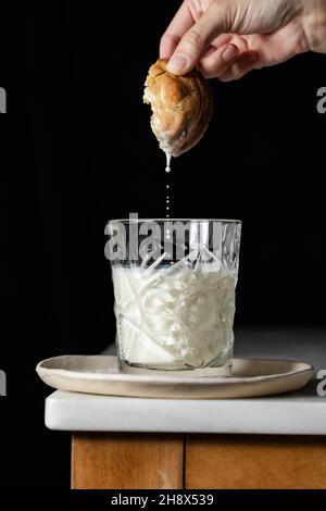 Crop anonyme Person taucht frischen süßen Cookie in ein Glas Milch auf der Ecke des Tisches vor schwarzem Hintergrund in der Küche platziert Stockfoto