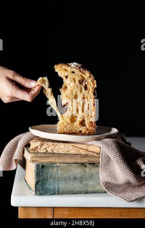 Anonyme Person, die vor dunklem Hintergrund einen geschnittenen Panettone isst, der mit einem Küchentuch auf dem Teller auf einem Stapel verwelkter Bücher liegt Stockfoto