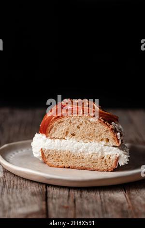 Appetitliche Scheibe Königskuchen, gefüllt mit weißer Creme auf dem Teller, der auf einem Holztisch vor schwarzem Hintergrund platziert ist Stockfoto