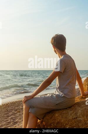 An einem sonnigen Tag sitzt ein junger Mann an der Küste. Einsamkeit und soziales Distanzkonzept. Gesichtslos. Selektiver Fokus. Stockfoto