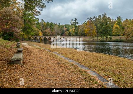 Rustikale Steinbänke gesäumt entlang des Wanderweges am Ufer des Sees mit einem Fischerdock und der gewölbten Brücke im Hintergrund am Kreuzkümmel Stockfoto