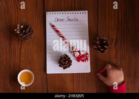 Lieber Weihnachtsbrief, Weihnachtskarte. Niedliches junges Kind, das einen Pullover trägt, einen Stift hält und auf einem weißen Blatt auf einem Holzhintergrund mit Kiefernkegel schreibt Stockfoto