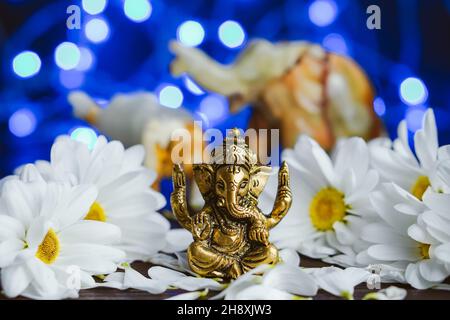 Goldener lord ganesha-Scluptur in Gänseblümchen auf blau beleuchtetem Hintergrund mit defokussierten Elefanten Stockfoto