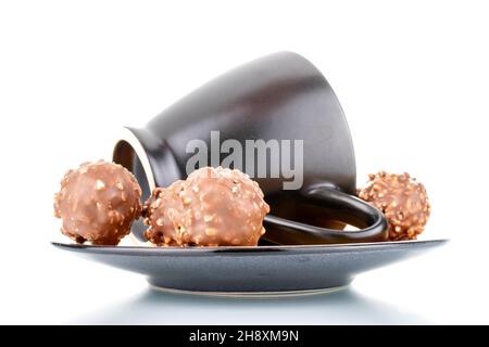 Drei süße Schokoladen-Bonbons mit Nüssen auf einer Keramikuntertasse mit einer Tasse, Nahaufnahme, isoliert auf Weiß. Stockfoto