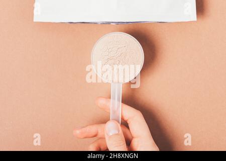 Löffel Proteinpulver mit Schokoladengeschmack in weiblicher Hand neben geöffneter Papiertüte auf braunem Hintergrund, Nahaufnahme, Draufsicht Stockfoto