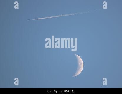 Fliegen Sie mich zum Mond oder in diesem Fall vorbei. Halbmond, ein Flugzeug und eine Dampfspur in klarem, blauem Himmel. Suffolk, Großbritannien Stockfoto