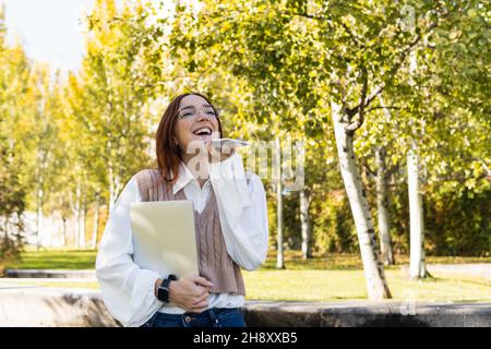 Lächelnde Frau mit einer Brille, die eine Audionachricht aufzeichnet oder die Spracherkennung verwendet, während sie ihren Laptop im Park hält. Junge Geschäftsfrau im Park Stockfoto