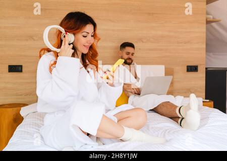 Die junge, frohhaarige Frau im Bademantel hört Musik mit Handy und Kopfhörer, während sie auf dem Bett sitzt Stockfoto