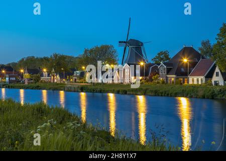 Typisches traditionelles holländisches Dorf mit Bauernhöfen und Windmühle entlang Kanal in der Dämmerung, Alkmaar, Noord Holland, Niederlande. Stockfoto