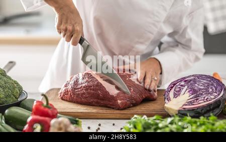 Die Köchin schneidet rohes Rindfleisch auf einem Holzbrett, das von Gemüse umgeben ist. Stockfoto