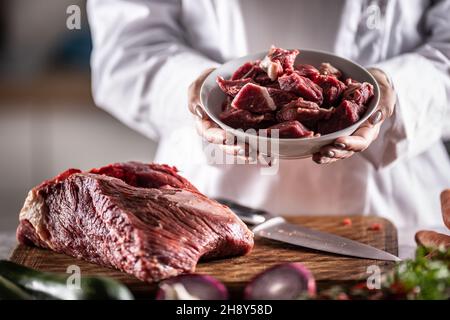 Kleine Stücke Rindfleisch oder Schweinefleisch zum Eintopf in einer Schüssel neben einem großen Stück rotem Fleisch auf einem Schneidebrett. Stockfoto