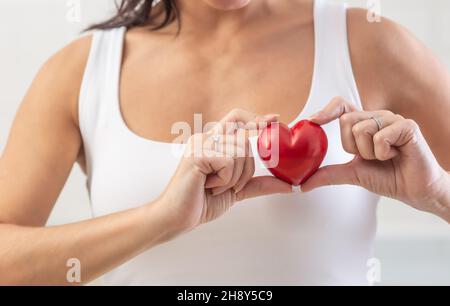 Die Frau hält ein kleines rotes Herz über ihrer Brust in dem Bereich, in dem das wahre Herz als Symbol für die Prävention von Herzerkrankungen steht. Stockfoto