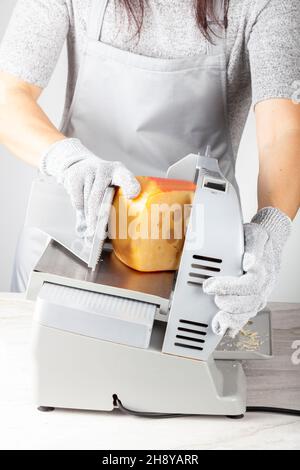 Ein Koch schneide einen großen Teil schweizer Käse mit einem elektrischen Deli-Slicer. Sie trägt beim Betrieb des Geräts schützende, schnittfeste Handschuhe Stockfoto