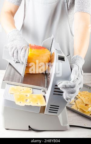 Ein Koch schneide einen großen Teil schweizer Käse mit einem elektrischen Deli-Slicer. Sie trägt beim Betrieb des Geräts schützende, schnittfeste Handschuhe Stockfoto